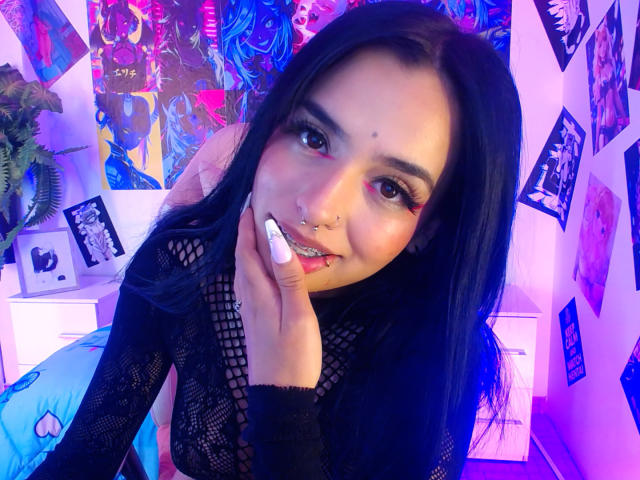 AtheneaOwenss with long black hair, pink eyeshadow, nose ring, and lip piercing, wearing black fishnet top, holding white nail, smiling at camera. Blue and pink neon lighting.
