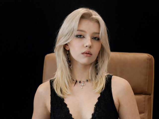 A photograph of SelenaIris with blonde hair and light skin, wearing a black lace sleeveless dress and a black necklace with a small cross pendant. She has green eyes and a neutral expression. She is seated in a tan leather chair against a black background. Her hair is straight and falls to her shoulders. She has small earrings in both ears. The lighting is even, highlighting her facial features and dress texture. The image is focused on her upper body.