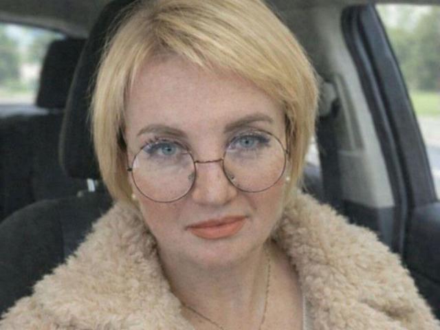 RenataDaster has a fair complexion with short, blonde hair and blue eyes. She wears round, metal-framed glasses and has light pink lipstick. She's dressed in a beige, fuzzy coat. The image is a close-up, focusing on her face from the chest up. The subtle texture of the coat contrasts with her smooth skin. Her expression is neutral, and the background is blurred, highlighting her as the central subject.