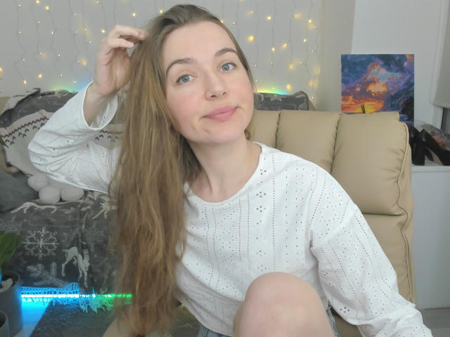 MonaTrio has long blonde hair, blue eyes, and light skin, wearing a white dotted long-sleeve shirt, sitting on a beige couch, smiling, with her hand in her hair.