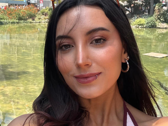 El rostro de CarlaFranco, cabello castaño oscuro, ojos color marrón claro, piel rosada, maquillaje sutil, labial rojo, arete pequeño de aro, top de tirantes color burdeos, ligera sonrisa, labio inferior perforado..