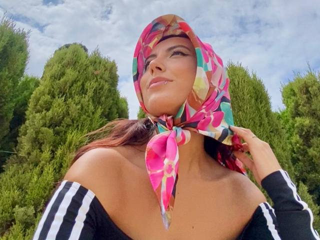Carla Franco, con una bufanda colorida, una blusa negra con un hombro descubierto y cabello castaño, mirando hacia arriba con una ligera sonrisa, de pie frente a árboles verdes bajo un cielo nublado..