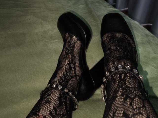 Os pés de Dreia, cobertos por renda preta, estão em saltos pretos brilhantes com detalhes prateados, com os tornozelos cruzados, sobre um espalhador verde. As pernas estão levemente bronzeadas, e os sapatos são brilhantes..