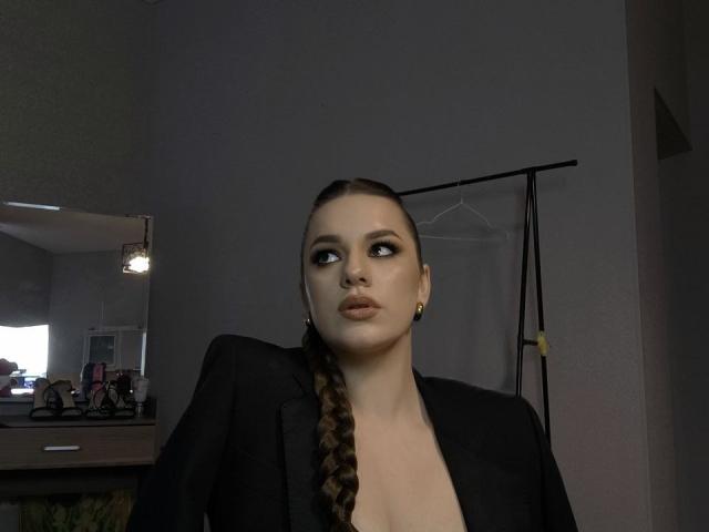 Dreia with a braided ponytail, wearing a black blazer, looking up with pouty lips, small gold earrings, and a slightly open top showing cleavage. Dimly lit room.