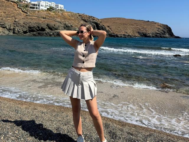 AnniaAnn stands on a pebbly beach, wearing a pink lace top and white pleated skirt, with hands behind her head, sunglasses on, and a sunny, clear blue ocean behind.