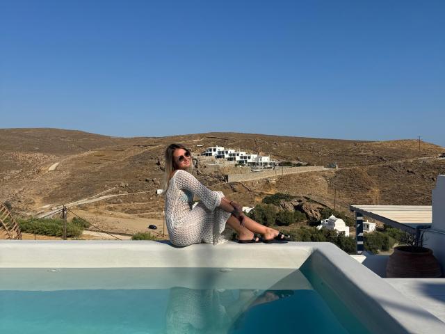 AnniaAnn, sitting on a pool's edge, wearing a white, sheer dress, sunbathing with her ass up, blue sky, brown hills, and white buildings in the background.