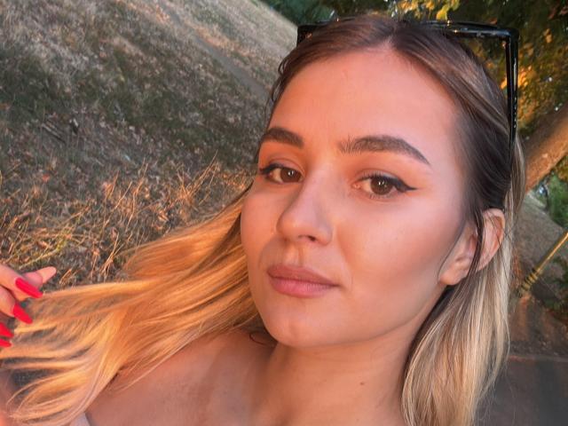 AnniaAnn has blonde hair, brown eyes, and is wearing black sunglasses. She's sporting red nails and is looking at the camera with a subtle smile.