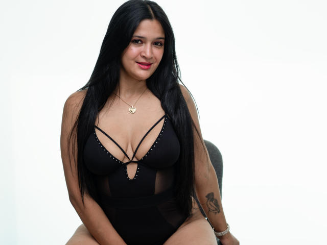 KiaraSienna with long black hair, wearing a black strappy lingerie that reveals her tits, sitting with a small tattoo on her right arm, smiling.