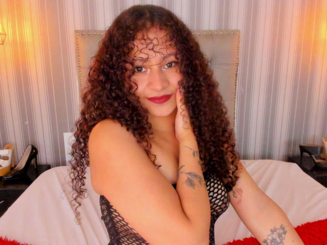 SofiaPassionne has curly, dark brown hair and fair skin. She's wearing a black fishnet top, revealing her cleavage. She has tattoos on her left wrist and right forearm. She's sitting on a bed with white sheets, a red blanket, and a beige, studded headboard behind her. Her right hand touches her face, and she's smiling with red lipstick. The room has warm, striped wallpaper and two wall lamps.