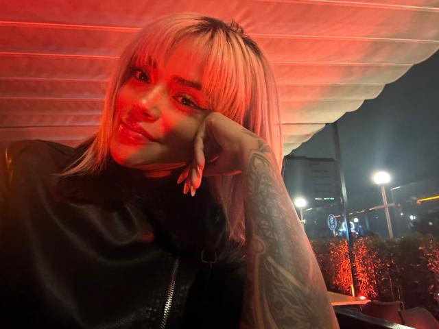 MajoJones with pink blonde hair, glossy red lips, black leather jacket, tattooed arm, resting chin on hand, red light on face, night scene, urban setting, sultry expression.