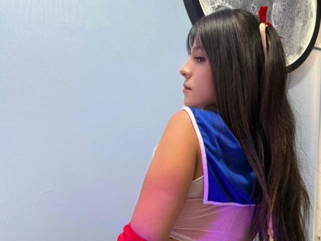 RilleyFoyx with long black hair, blue and white top, red arm warmers, looking over shoulder, white wall, and circular pattern in background. Boobs visible from side.