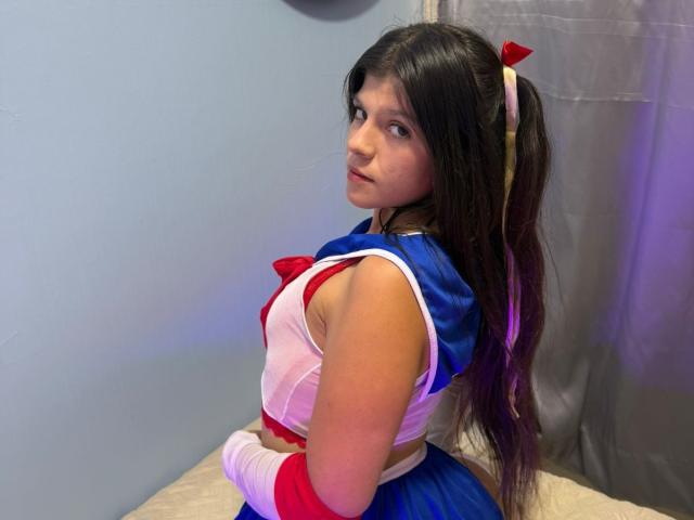 RilleyFoyx with long black hair in a blue and red cosplay outfit, looking back with a serious expression, showing white bandaged wrist, and small white-haired red bow.