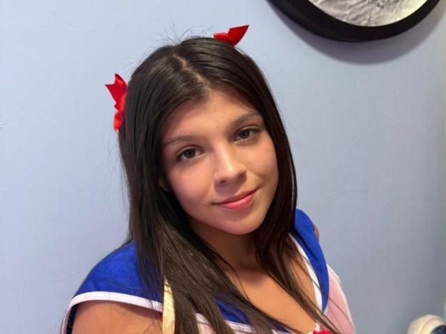 RilleyFoyx has long brown hair, red hairbows, and a cute smile, wearing a blue and white sailor-style top, with small, cute tits, and a slightly visible cleavage.
