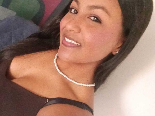 NaomiRios with medium brown skin, long black hair, smiling, wearing a black top, pearl necklace, and pearl earrings, showing her tits and white teeth.