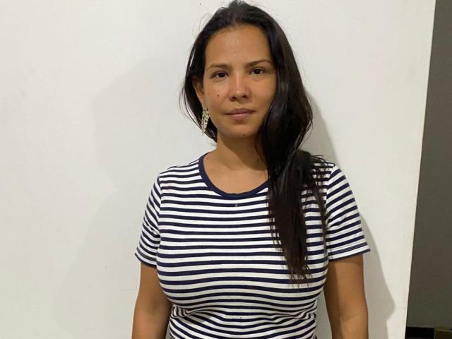 AfroditaSecret with medium tits, brown skin, long black hair, wearing a striped navy and white shirt, standing against a white wall, looking slightly to the side.