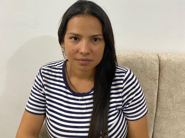 AfroditaSecret has long dark hair, brown eyes, and a small mole on her right cheek. She's wearing a navy and white striped shirt, sitting on a beige chair.