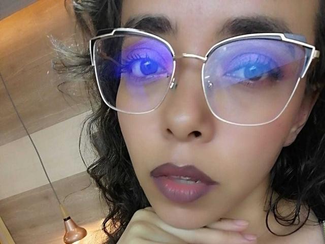 BarbaraLeythonn with blue neon eyes in oversized, reflective glasses, has curly black hair, purple lipstick, and a serious expression, with her hand near her chin.