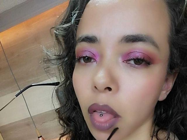 Barbara Leythonn tem sombra rosa, olhos castanhos, cabelo preto encaracolado, um piercing no nariz e uma língua com piercing, com uma bolinha preta, olhando para a câmera com os lábios roxos entreabertos..