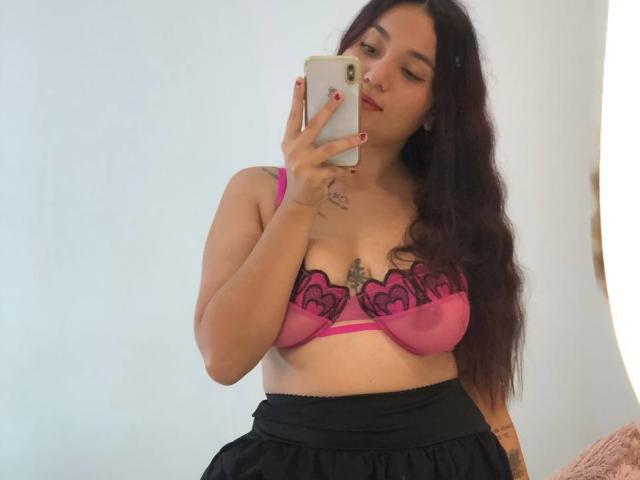 IvyJoness with long dark hair, winking, holding a phone, wearing a pink bra with black lace, black skirt, tattoos on chest and arm, taking a mirror selfie.