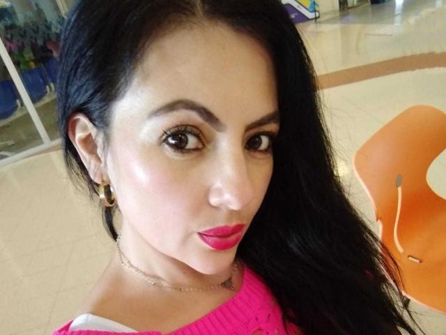 JakyKidman with shiny skin, black hair, brown eyes, pink lipstick, gold hoop earrings, and a pink sweater, looking at the camera with a slight smile.