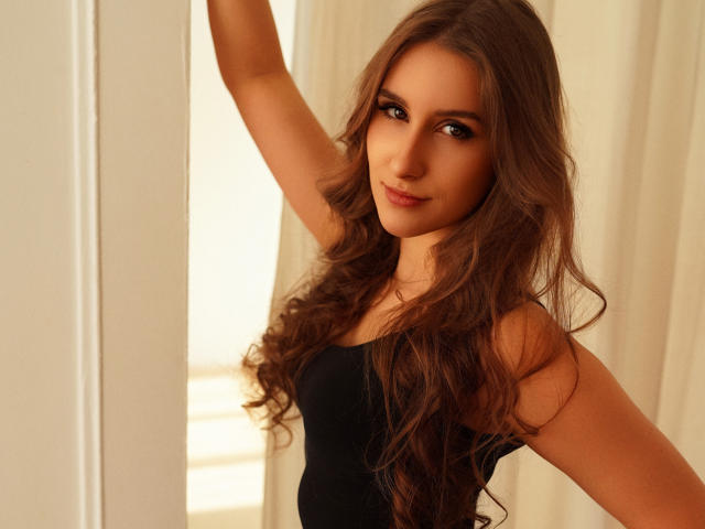 VanessaTrue with long brown wavy hair, wearing a black tank top, raising her right arm, smiling with brown eyes, and a cute face. Her tits are small and she's hot.