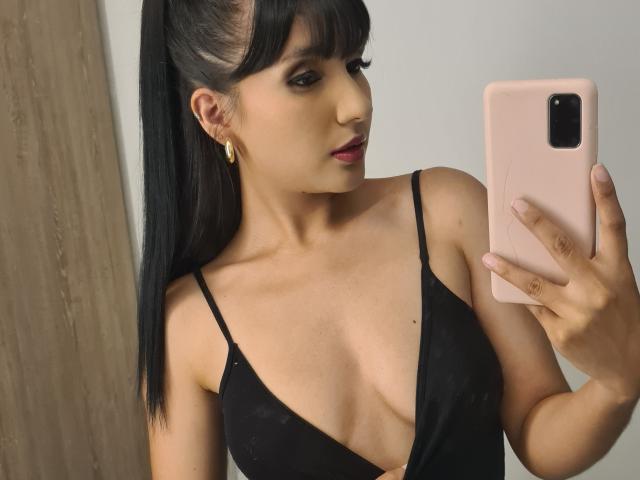 NaylaGrey with black hair in a ponytail, wearing a low-cut black dress, showing her tits, holding a pink phone, gold earrings, and pink lipstick, taking a mirror selfie.