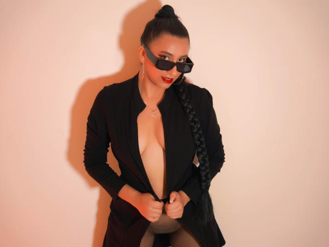 Catalina Rivero, con cabello negro trenzado, usando gafas de sol negras, una chaqueta negra con un escote en V pronunciado, pantalones de malla, que dejan ver sus pechos, con lápiz labial rojo, y un anillo en el pecho..