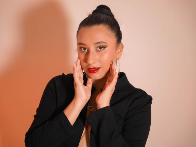 CatalinaRivero has dark hair in a bun, wearing red lipstick, a black jacket, and heart-shaped earrings, with hands touching her face, showing off her tits subtly.