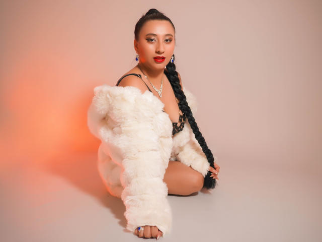 Catalina Rivero, agenouillée, porte un manteau blanc duveteux, un soutien-gorge noir et une longue tresse. Elle a du rouge à lèvres, des boucles d'oreilles et un collier. Ses seins sont visibles, et ses fesses sont légèrement dissimulées..