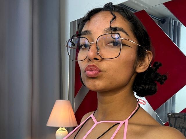 TangiiSoul with curly black hair, wearing glasses and pink halter top, pouting with pink lips, brown skin, small zit on cheek, standing next to a lit lamp.