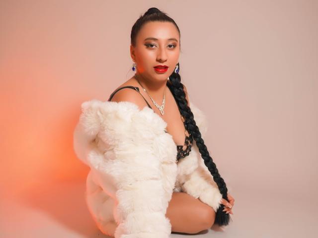CatalinaRivero with long black braid, wearing fluffy white coat, black lace lingerie, red lipstick, and diamond necklace, sitting, looking at camera, ass partially visible, sexy, confident.