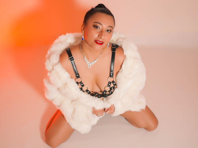 CatalinaRivero with dark skin, red lipstick, and blue earrings, kneeling, wearing a fluffy white coat, black strappy lingerie, and a diamond necklace, showcasing her large, rounded tits.