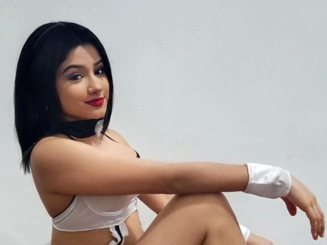 SuesseKlara-hot with black hair, red lipstick, white bra, white wrist cuffs, and black neck scarf, sitting, showing tits, and smiling at the camera.