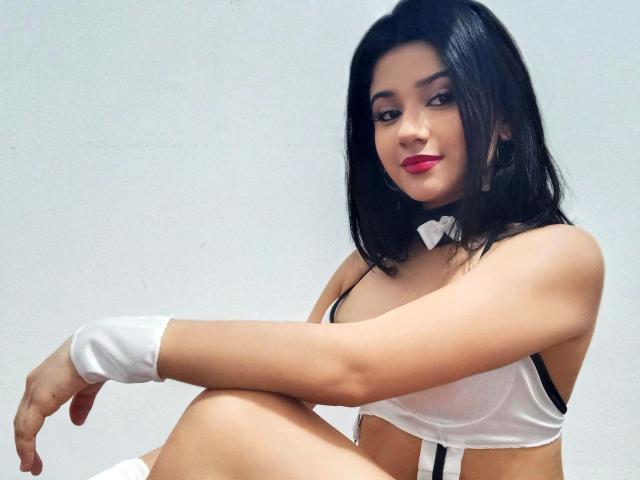 SuesseKlara-hot with black hair, red lipstick, white bra, black straps, white wrist cuffs, and white bowtie, sitting with knees up, showing her tits.