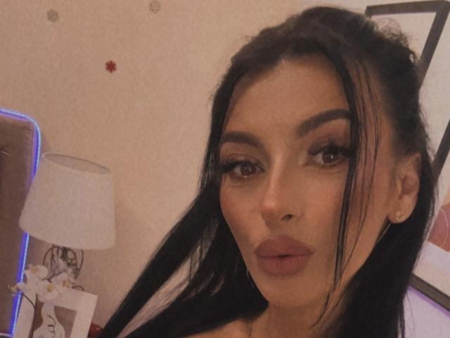 KinkyAmelie with long black hair, full eyebrows, and thick eyelashes, has pouty brown lips and a tan complexion, wearing small gold earrings, in a slightly blurred selfie.