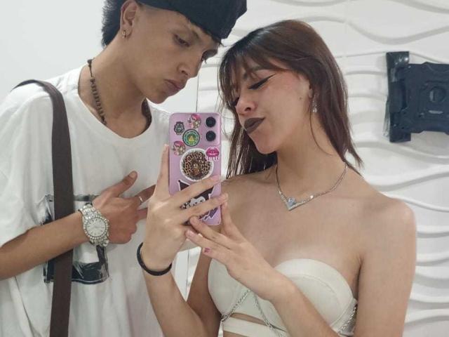 FeviceFabinLm with brown hair and dark lipstick, showing her phone with stickers, to FeviceFabinLm in a white shirt and black cap, standing by her. Her tits are visible.