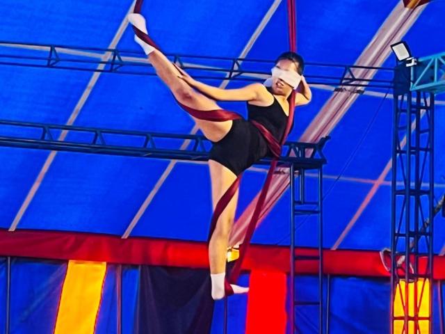 HattyMollie is suspended in mid-air, wearing a black leotard with red ribbons, doing a high leg kick. Her blindfolded face shows concentration. She has a cute ass.