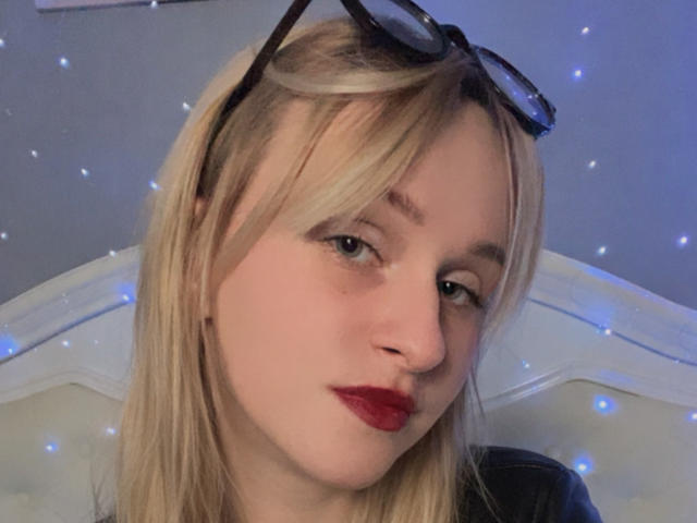 FlyFreedom has blonde hair, glasses on her head, red lipstick, and light skin. She's looking to the side with a slight smile, showing her nose and upper lip.