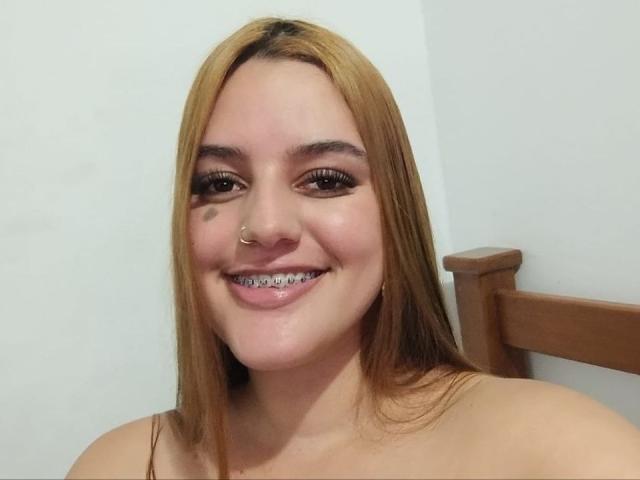 HaileyTims has light brown hair, a nose ring, a small black tattoo under her right eye, and a smile showing metal teeth. She's sitting with a wooden chair.