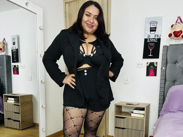 KarlaWild with black hair, medium tits, in a black blazer, black bra, high-waisted shorts, and patterned fishnet stockings, smiling confidently, standing in a bedroom.