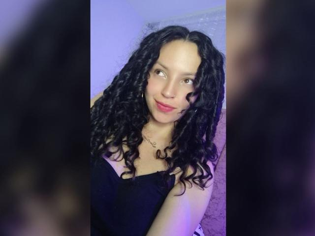 WildSelena69 with curly black hair, light brown skin, and pink lipstick, wearing a black dress, smiling softly, reflecting in a mirror. Boobs visible under dress, small to medium size.