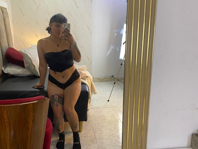 LilithVelvet, avec sa peau mate, un haut noir sans bretelles, des sous-vêtements noirs, un tatouage sur la cuisse gauche, et des chaussettes noires, prend un selfie devant un miroir, montrant ses fesses et son vagin..