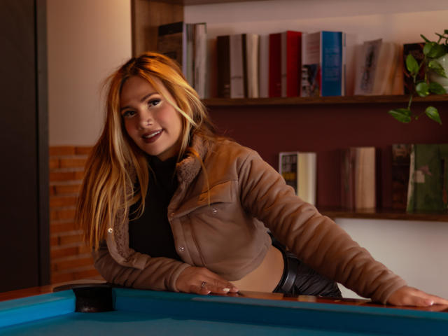 AzhunaStar, con lunghi capelli biondi, giacca marrone, top nero, appoggiata a un tavolo da biliardo blu, sorridente, che mostra il seno, e indossa pantaloni neri in una stanza poco illuminata..