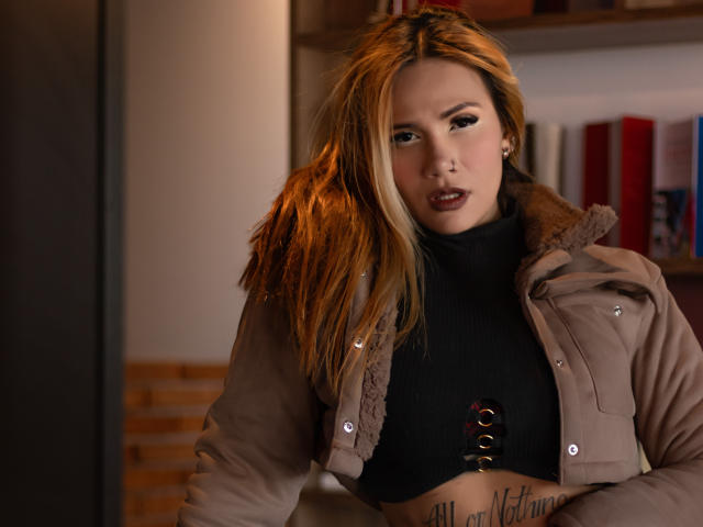 AzhunaStar with blonde hair, wearing a beige coat and black crop top with lace-up front, exposing her tits and lower belly tattoo, looking seductively at the camera.
