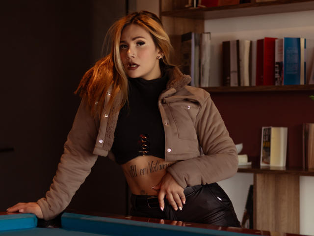 AzhunaStar stands in a dimly lit room, leaning on a blue pool table. She has long, blonde hair and wears a beige jacket over a black crop top with a tattoo of "Melissa Gellert" below her right breast. Her black leather pants are tight. She has a serious expression, dark makeup, and light skin. Shelves with books are in the background. The image has a realistic, photographic style.