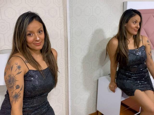 CynthiaMoore with long brown hair, wearing a sparkly black dress, showing off tattoos on her right arm, sitting with her ass on a white box, smiling. Reflection in mirror.