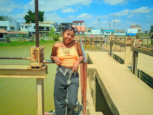 QueenBella69 with dark skin, orange top, gray pants, standing by a rusty metal bridge, wearing black sunglasses, smiling, hands on bridge, blue sky, green water, and colorful buildings.