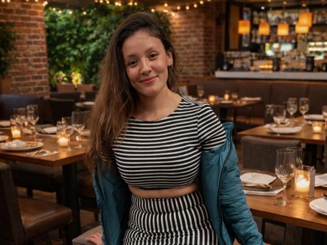 CynthiaMoore with long brown hair, smiling, wearing a striped black and white crop top and matching skirt, blue jacket off, sitting in a dimly lit restaurant with candles.