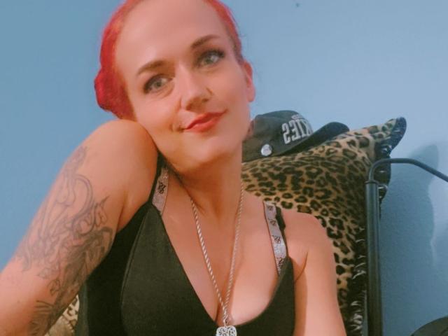 Red-haired HIMandHER with tattoos on her arm, black tank top, necklace, and subtle smile, sitting against leopard print pillow, blue wall background. Boobs visible, small to medium.