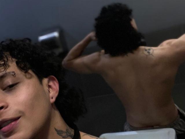 JayTaylor, avec ses cheveux noirs bouclés et son piercing au nez, torse nu, tatouage sur le haut du dos, bras levés, musclé, petite fesse, reflet dans un miroir, intérieur, arrière-plan légèrement flou..